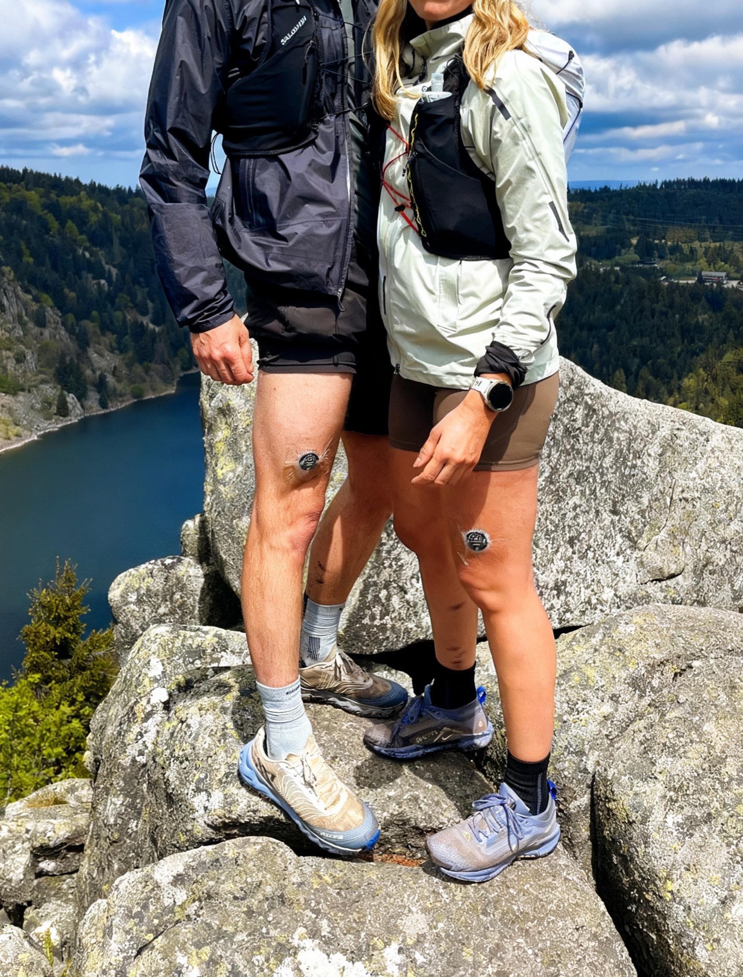 Two trail runners at a mountain summit, each wearing an AUSPRÁ sensor on their thigh